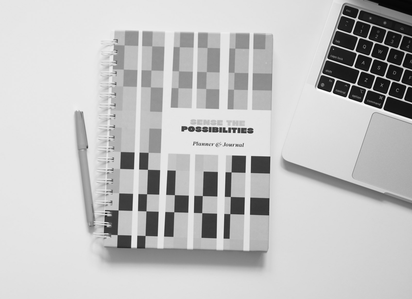 The Sense the Possibilities planner and journal, with a checkerboard cover, beside a pen and a laptop