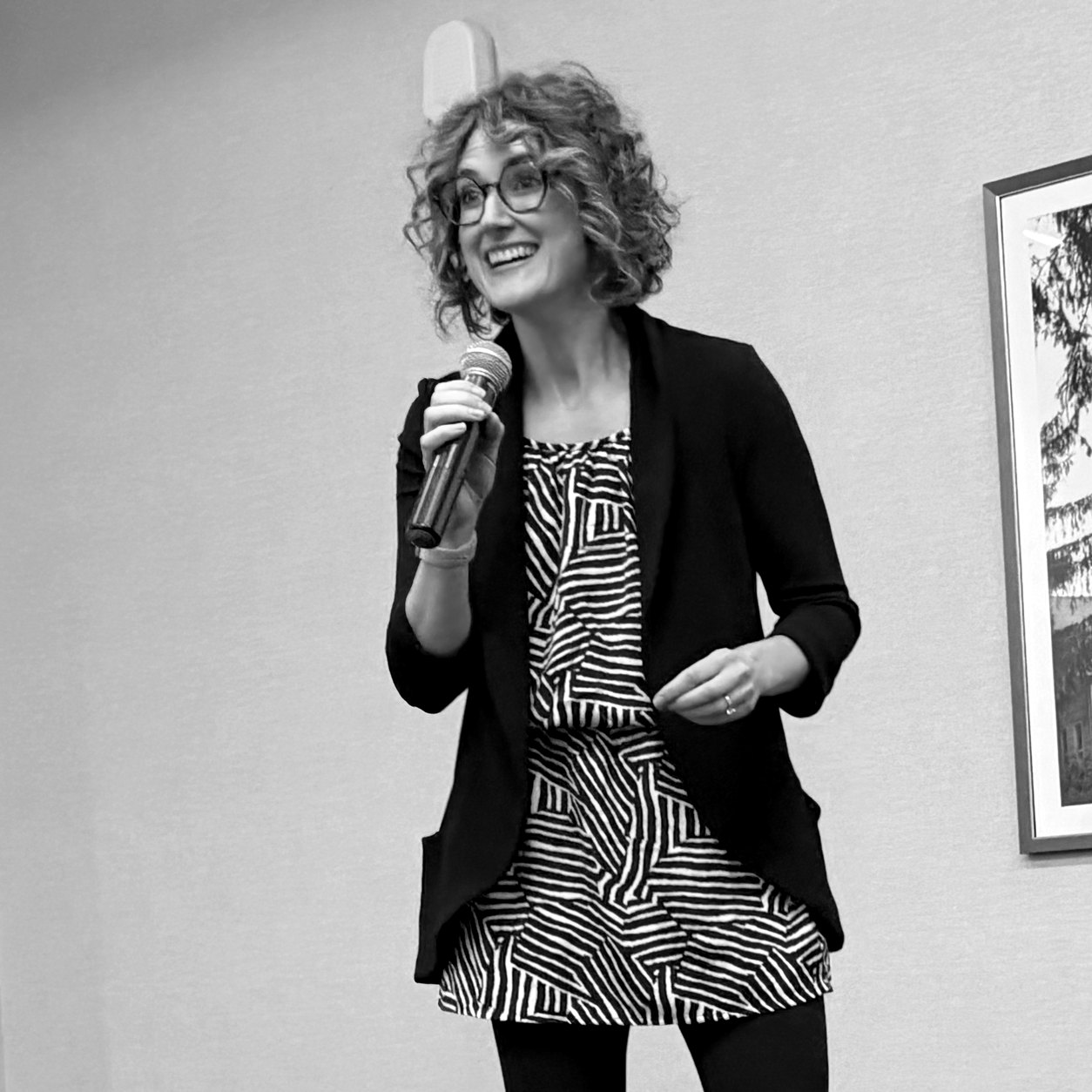 Danielle McGeough speaking into a microphone on a stage, smiling, in glasses and a black blazer