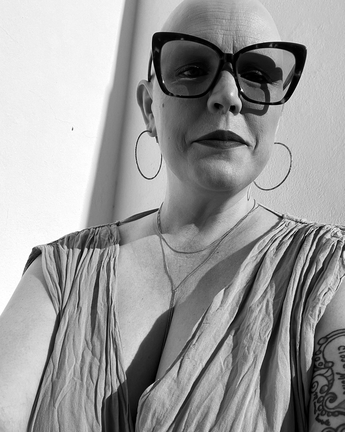 Laura Cole in large cat-eye glasses and hoop earrings, photographed against a sunlit white wall