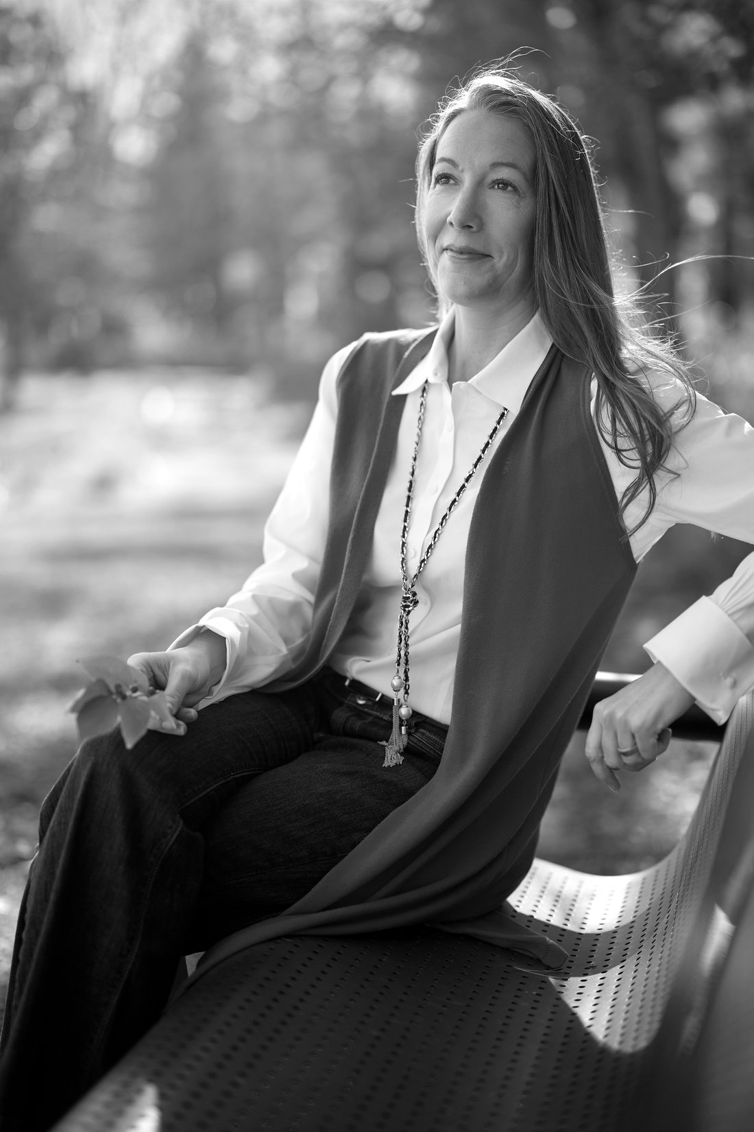 Monica Phillips seated on a park bench, a scarf over a white shirt, holding a sprig of leaves