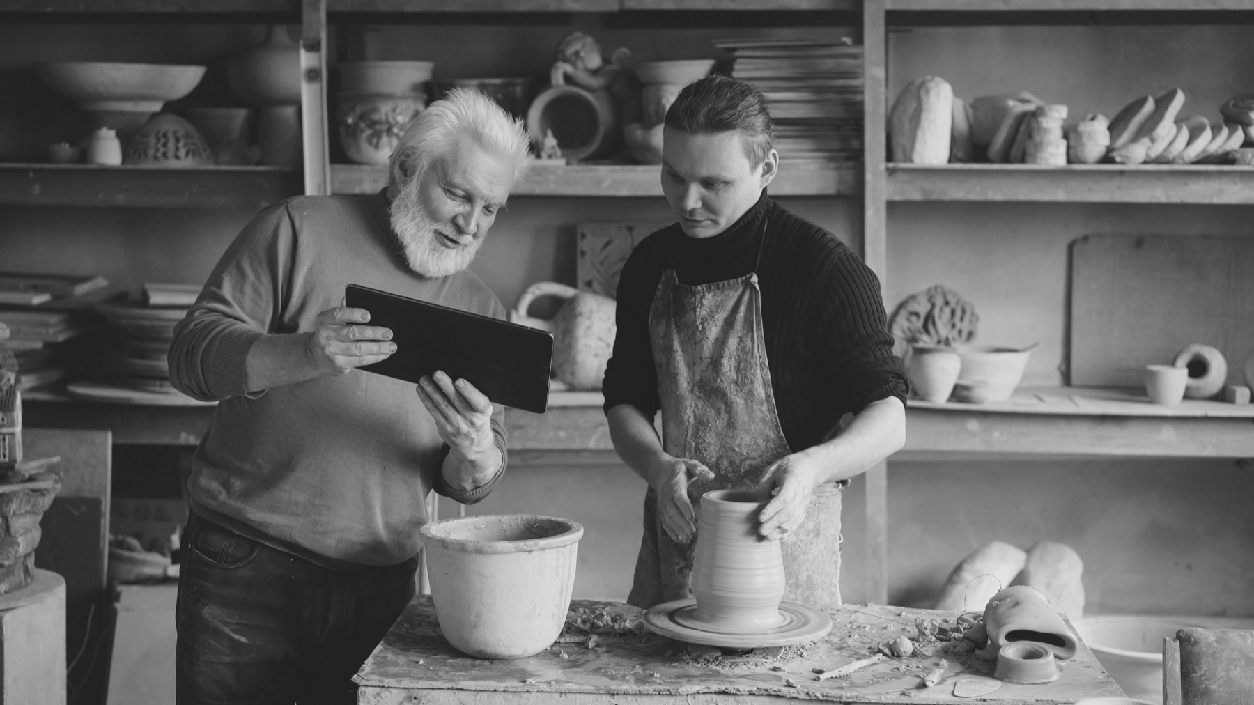 An older potter holding a tablet while a young apprentice shapes a vase at the wheel